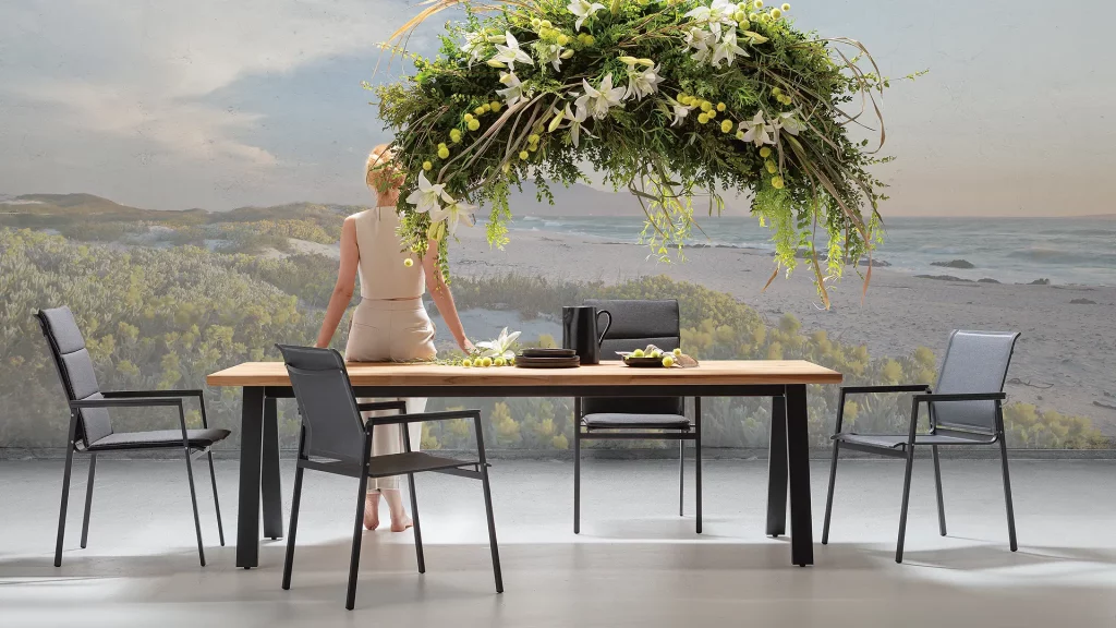 Moderne Outdoor-Essgruppe mit rechteckigem Holztisch und vier schwarzen Stühlen im Gewebe-Design, arrangiert vor einer malerischen Strandkulisse am Meer gelegen. Stilvolle Gartenmöbel schaffen eine elegante und zugleich naturnahe Atmosphäre für Terrasse, Balkon oder Garten.