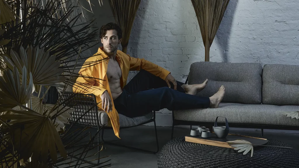 Moderner Outdoor-Loungebereich mit grauem Sofa, schwarzem Geflecht-Hocker als Couchtisch und Teeservice, in einem stilvollen Setting mit Naturdeko und Palmenblättern. Eine Person in gelber Jacke sitzt entspannt auf einem Stuhl. Das hochwertige Gartenmöbel-Arrangement eignet sich ideal für Terrasse, Balkon oder Garten